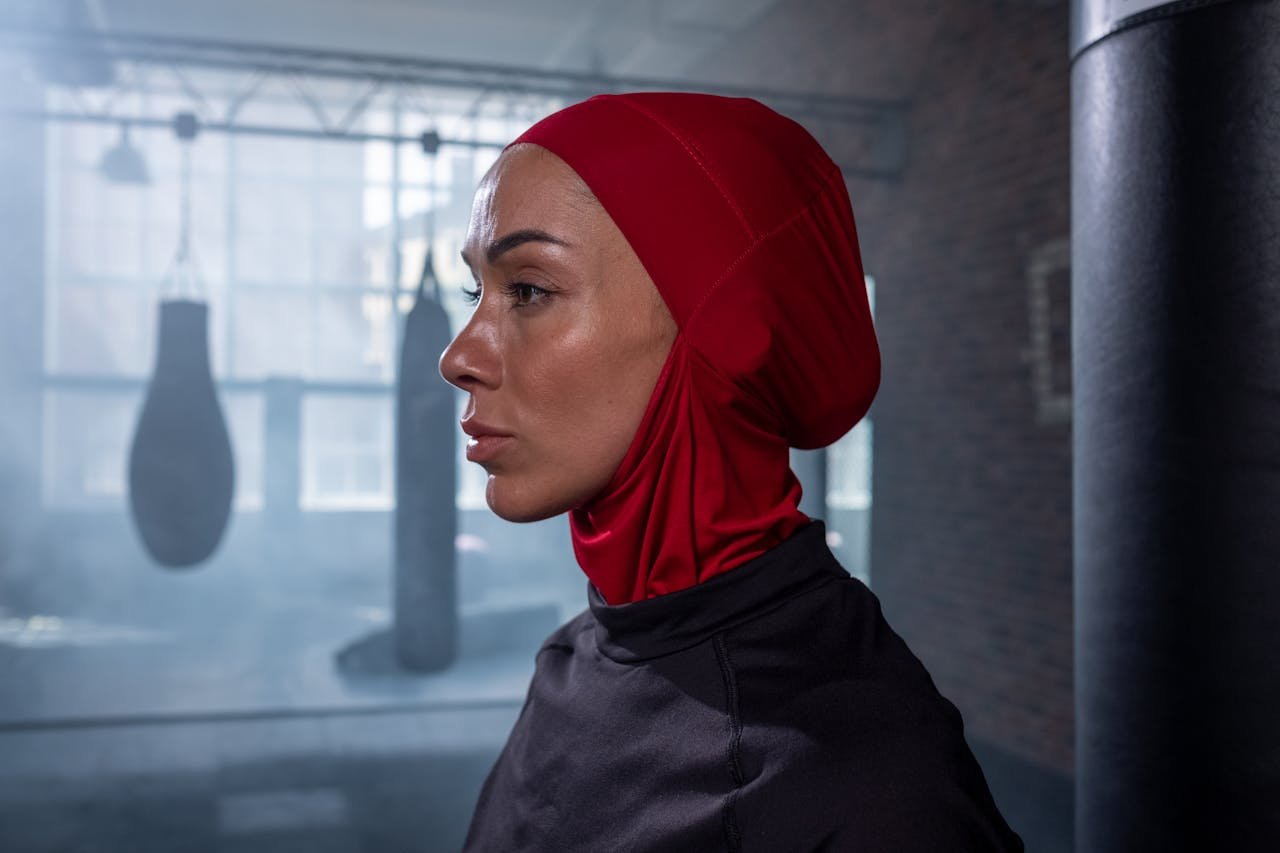 Side profile portrait of a female boxer in a red hijab at the gym.